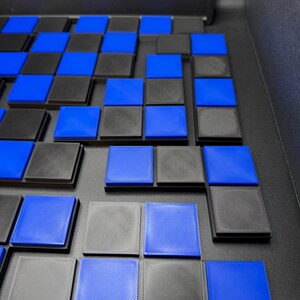 May include: A partially assembled board game with a checkerboard pattern. The squares alternate between black and blue. The game pieces are square and have a raised edge. The board is black.