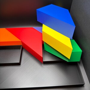 May include: A set of geometric tangram-style blocks in vibrant blue, yellow, green, red, and orange colors. The pieces are arranged in a stack and alongside a flat, black rectangular tray with multiple compartments for organizing the shapes.