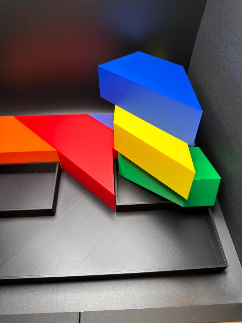 May include: A set of geometric tangram-style blocks in vibrant blue, yellow, green, red, and orange colors. The pieces are arranged in a stack and alongside a flat, black rectangular tray with multiple compartments for organizing the shapes.