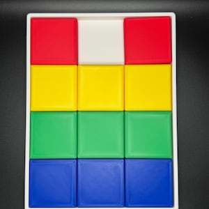 May include: A white tray with nine square compartments, each filled with a different colored block. The blocks are red, yellow, green, and blue.