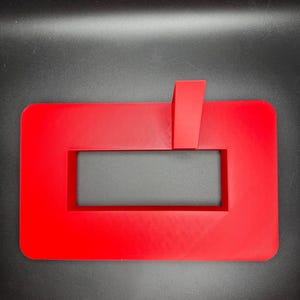 May include: A red plastic stand with a rectangular cutout and a small upright piece on top. The stand is designed to hold a phone or tablet.