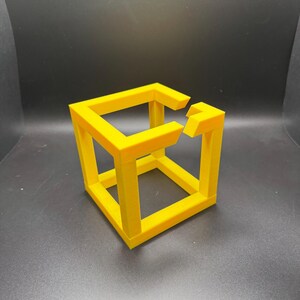 May include: A bright yellow impossible cube sculpture, a 3D printed optical illusion. The cube appears to defy the laws of physics with its interconnected, yet disjointed, structure. The object is set against a dark background.