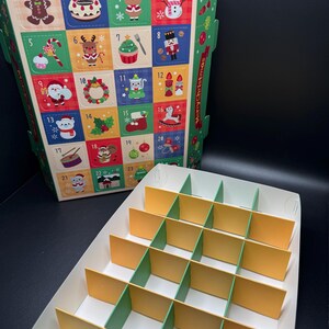 May include: A Christmas advent calendar box with colorful squares featuring festive illustrations and numbers. The box is open, revealing a compartmented interior for treats. The words "Merry Christmas" are printed on the side.