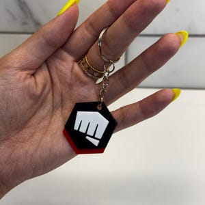 May include: Black, red, and white hexagon-shaped keychain with a fist graphic. The keychain is attached to a silver keyring. The design features a black background with a red border and a white fist.
