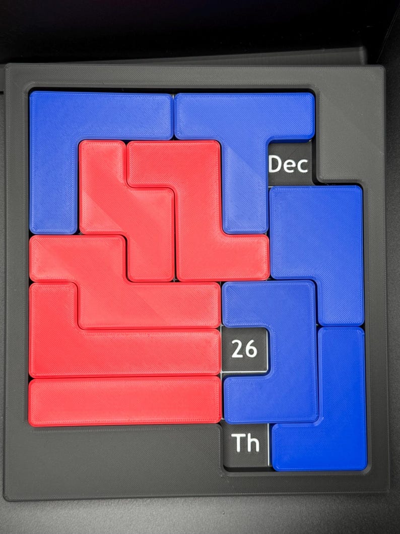 K&ouml;nnte beinhalten: Ein rotes und blaues Puzzle mit dem Datum 26. Dezember darauf. Das Puzzle ist aus Kunststoff und hat einen schwarzen Rahmen.