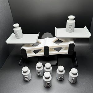 May include: A white plastic balance scale with two platforms. The scale is tipped to the right side. A white plastic weight with the number 3 is on the left platform and a white plastic weight with the number 5 is on the right platform. There are seven additional white plastic weights with the numbers 1, 2, 3, 4, 5, 6, and 7 on the surface below the scale.