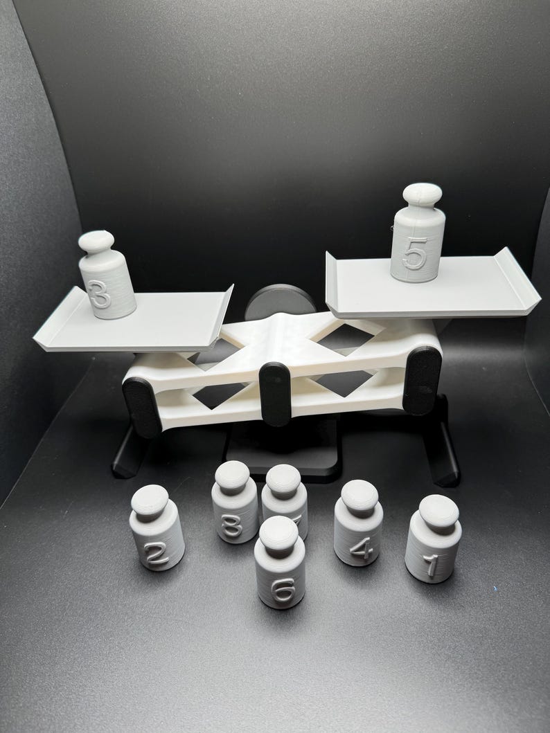 May include: A white plastic balance scale with two platforms. The scale is tipped to the right side. A white plastic weight with the number 3 is on the left platform and a white plastic weight with the number 5 is on the right platform. There are seven additional white plastic weights with the numbers 1, 2, 3, 4, 5, 6, and 7 on the surface below the scale.