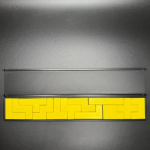 May include: A set of yellow wooden puzzle pieces in a black box. The pieces are arranged in a pattern that resembles a maze.