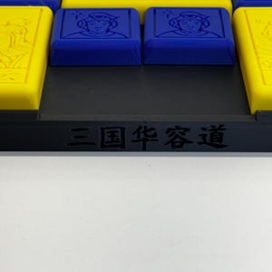 May include: A close-up of a game board with a black frame and the text "三国 策略" in white. The board has a grid of squares with different colored tiles, including yellow, red, and blue. The blue tiles have images of people on them.