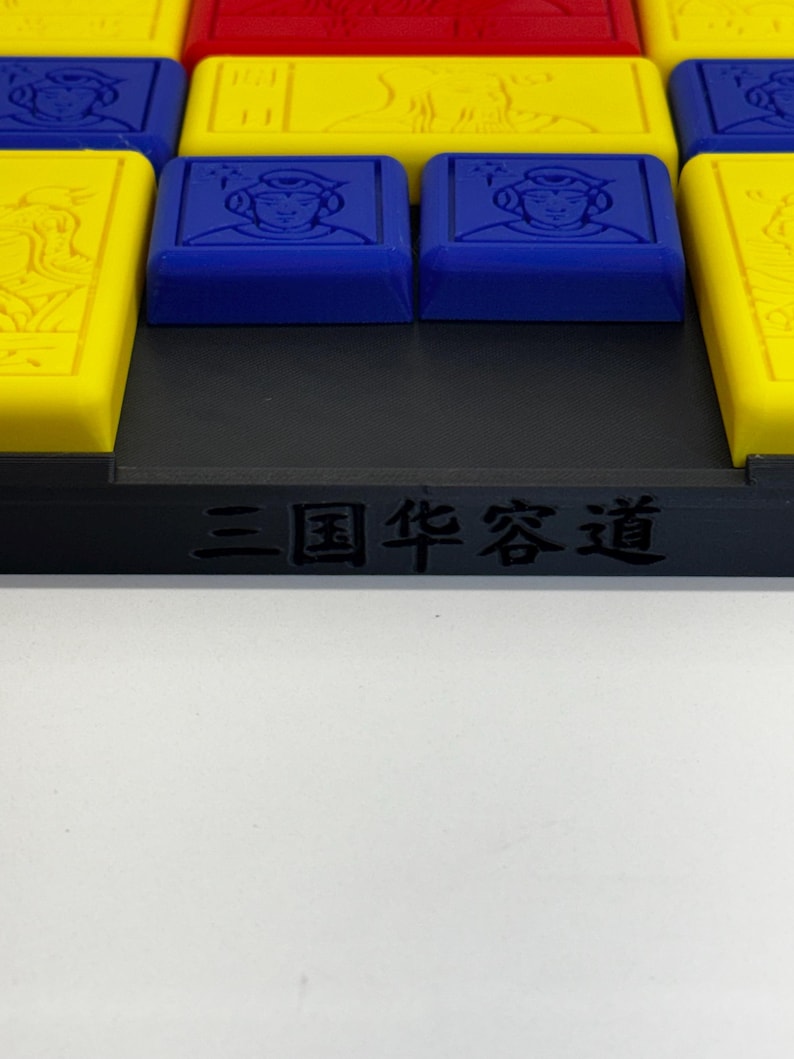 May include: A close-up of a game board with a black frame and the text "三国 策略" in white. The board has a grid of squares with different colored tiles, including yellow, red, and blue. The blue tiles have images of people on them.