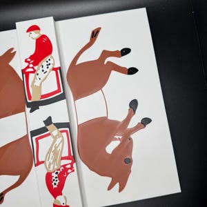 May include: A set of three white wooden tiles with a red, white, and black illustration of a jockey riding a brown horse. The horse is in a jumping position with its legs in the air.