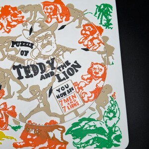 May include: A white puzzle featuring a circular design with the text "Puzzle of Teddy and the Lion." The design includes illustrations of lions, men, and foliage in orange, green, and tan colors. The text "You Now See 7 Men and 7 Lions" is also present.