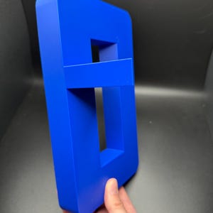 May include: A bright blue plastic letter 'B' with a hollow center. The letter is standing upright on a dark surface.