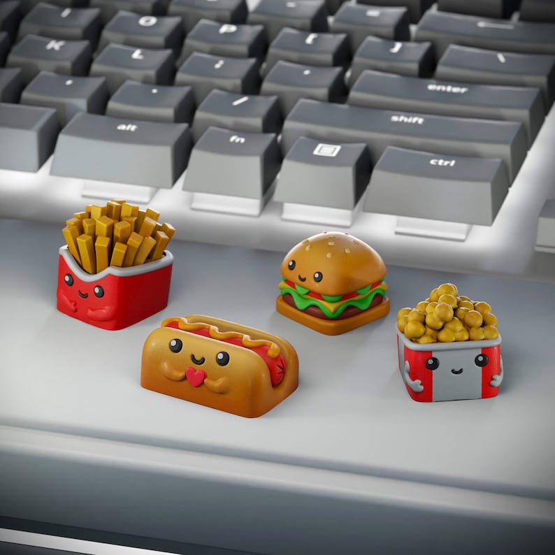 May include: Four cute cartoon food keycaps: a red box of french fries, a hot dog, a hamburger, and a red box of popcorn. The keycaps are on a gray keyboard.