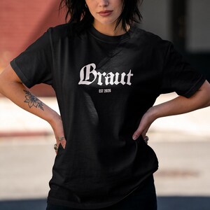 Könnte beinhalten: Ein schwarzes kurzärmeliges T-Shirt mit Rundhalsausschnitt, das den weißen Schriftzug Braut in einer gotischen Schriftart mittig auf der Brust zeigt, mit dem kleineren weißen Text EST 2026 direkt darunter in einer serifenlosen Schrift.