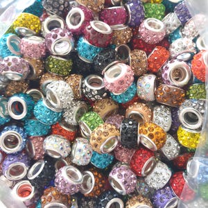 May include: A large assortment of colourful, round, rhinestone-encrusted beads. The beads are all different colours, including red, blue, green, purple, and pink. They are all about the same size and have a silver metal ring in the centre.