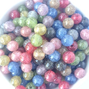 Op de afbeelding: Een verzameling kleurrijke, glinsterende kralen in verschillende kleuren, waaronder roze, blauw, groen en goud. De ronde kralen zijn gemaakt van een doorschijnend materiaal, met glitter erin. De kralen liggen in een witte kom.