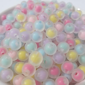 Puede incluir: Un bol lleno de cuentas translúcidas de colores pastel. Las cuentas son redondas y tienen un acabado mate. Los colores incluyen rosa, azul, amarillo y morado.
