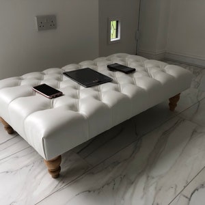 May include: A white tufted ottoman with four wooden legs. Three electronic devices are placed on top of the ottoman, including a tablet and two smartphones.