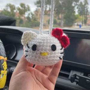 May include: White crocheted Hello Kitty car charm with a red bow. The charm is hanging from a white string.