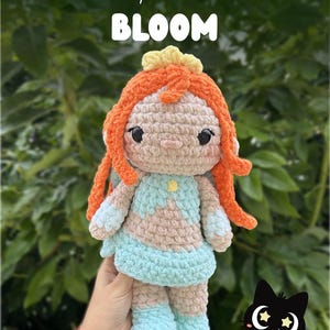 Op de afbeelding: Een gehaakte pop met oranje haar, een lichtblauwe jurk en een gele bloemenkroon. De pop heeft zwarte ogen en wordt vastgehouden voor een groene, bladerrijke achtergrond. De tekst "crochet pattern release BLOOM" staat bovenaan.