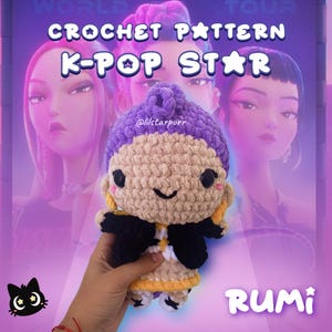 Op de afbeelding: Gehaakte pop met een paarse hoed, een bruin gezicht en een zwarte jas. De pop heeft een lachend gezicht en wordt in een hand gehouden. De tekst op de afbeelding luidt "CROCHET PATTERN K-POP STAR" en "RUMI".