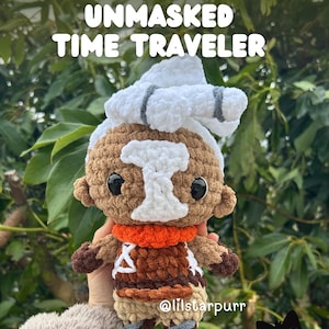 May include: A crochet doll featuring a tan face, white hair, and a white mask-like patch over the eyes. The doll wears a brown tunic with white accents, an orange scarf, and dark brown boots. Text reads: Crochet patterm release UNMASKED TIME TRAVELER.