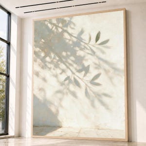 Puede incluir: Una gran pieza de arte mural rectangular en un marco de madera clara que presenta una superficie texturizada de color blanco roto con la sombra suave y gris oscuro de una rama de olivo proyectada, creando un efecto visual de luz y profundidad.