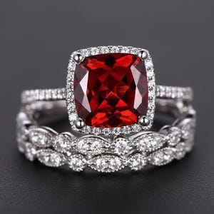 May include: A two-piece ring set featuring a large, square-cut, red gemstone surrounded by a halo of smaller, clear stones. The band is also adorned with clear stones. The ring set is displayed against a dark background.
