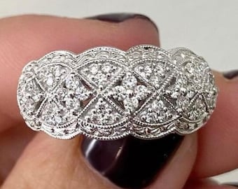 May include: A detailed, silver-toned ring with a repeating pattern of diamond-like stones. The ring features an intricate design with a series of diamond-shaped sections, each filled with smaller stones. The ring is held by a person.