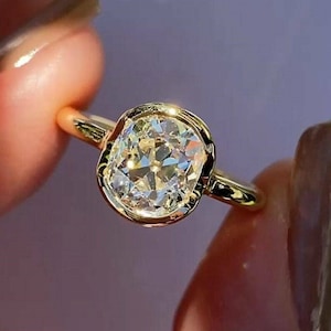 May include: A gold ring featuring a large, oval-cut diamond. The diamond is set in a bezel setting, and the band is a simple, polished gold. The ring is held up by a person's fingers, showcasing the ring's sparkle and design.