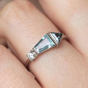 May include: A silver ring with a light blue aquamarine gemstone and two smaller diamonds. The aquamarine is a long, rectangular shape with a pointed end. The diamonds are round and set on either side of the aquamarine.