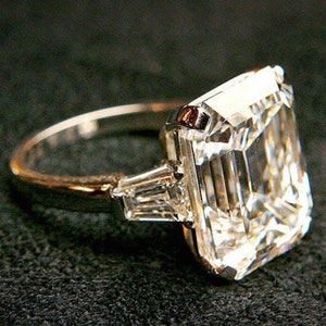 May include: A large, clear, emerald-cut diamond ring set in a platinum band. The diamond is surrounded by two smaller, baguette-cut diamonds on either side.