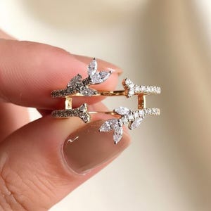 May include: A gold-colored ring with a leaf design. The ring features clear, sparkling stones and is held by a person. The ring has a unique design with two bands connected by vertical bars.