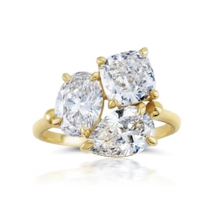 May include: A gold ring featuring three large, sparkling diamonds in oval, cushion, and pear shapes. The diamonds are set in a cluster design, with a polished gold band. The ring is displayed against a white background.
