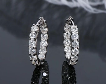 May include: A pair of silver hoop earrings, each encrusted with a row of sparkling, round-cut white gemstones. The earrings are displayed against a dark, reflective surface, highlighting their brilliance.