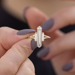 May include: A gold ring with a vertical rectangular design, set with three clear, rectangular-cut stones. The ring has a slender band. The ring is held between two hands with painted nails.