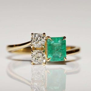 May include: A gold ring featuring two clear, square-cut diamonds and a large, emerald-cut green emerald. The ring is set against a white background, with a reflection visible below.