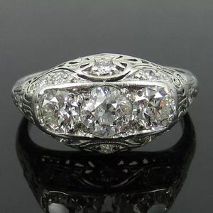 May include: A silver ring with three large, round diamonds set in the center, flanked by smaller diamonds. The ring has an intricate, engraved design on the band and around the stones, showcasing a vintage or antique style.
