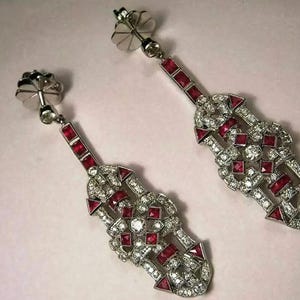 Könnte beinhalten: Ein Paar Ohrhänger im Art-Deco-Stil. Die Ohrringe haben eine silberfarbene Metalleinfassung mit geometrischem Design. Sie sind mit kleinen, runden und quadratischen roten Edelsteinen und klaren, funkelnden Steinen verziert.