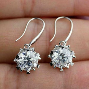 May include: A pair of silver drop earrings with large, round, clear gemstones. The gemstones are surrounded by a decorative, starburst-like setting. The earrings have a simple hook closure.