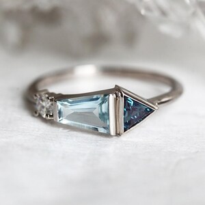 May include: A silver ring with a light blue aquamarine gemstone and a dark blue triangular gemstone. The ring has a delicate design with small diamonds on the band.