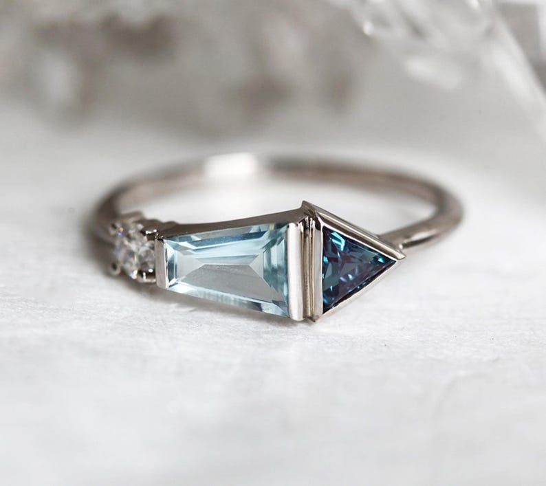 May include: A silver ring with a light blue aquamarine gemstone and a dark blue triangular gemstone. The ring has a delicate design with small diamonds on the band.
