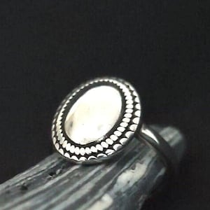 May include: A silver ring with an oval-shaped top. The top of the ring has a textured, beaded design around the edge.