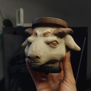 Peut inclure: Un pot en céramique en forme de tête de chèvre avec quatre yeux. Le pot est peint en blanc avec des accents bruns et a un dessus en bois.