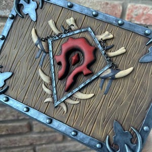 Op de afbeelding: Een houten schild met een zilveren metalen frame en accenten. Het schild heeft een rood embleem in het midden, omringd door botachtige slagtanden en een ketting. Het hout heeft een getextureerd nerfpatroon.