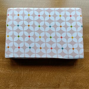 May include: A rectangular box with a floral design. The pattern shows white flower shapes on a pale pink backdrop, with colourful dots at the centre of each flower. The box is likely for storage or as a decorative item.