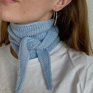 May include: A light blue knitted scarf with a knotted design. The scarf is worn around the neck and has two long ends that hang down.