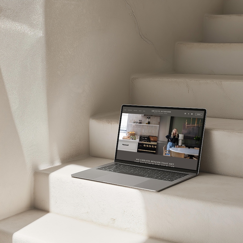 May include: A silver laptop displaying a website on white stairs. The website features a kitchen design with a person in the background. The text on the website reads "WE CREATE SPACES THAT SET THE STAGE FOR YOUR BEST LIFE."
