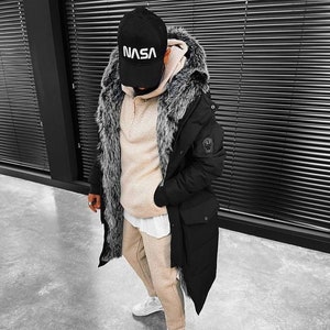 May include: A black parka with a faux fur-lined hood, a beige herringbone textured hoodie, a white undershirt, beige herringbone joggers, white sneakers, and a black baseball cap with the white text NASA embroidered on the front panel.